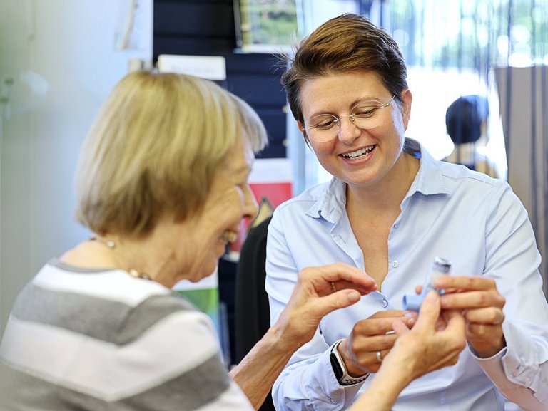 Apothekenmitarbeiterin zeigt einer älteren Kundin die Handhabung eines Inhalators; beide lächeln und betrachten das Gerät gemeinsam.
