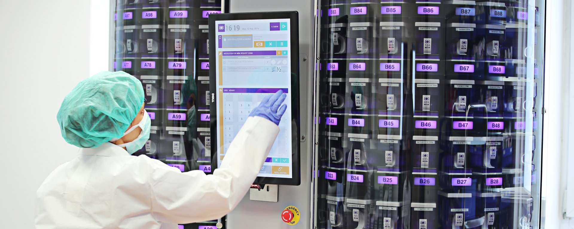 Mitarbeiterin in Schutzkleidung bedient per Touchscreen eine automatische Verblisterungsmaschine mit vielen Medikamentenkassetten.