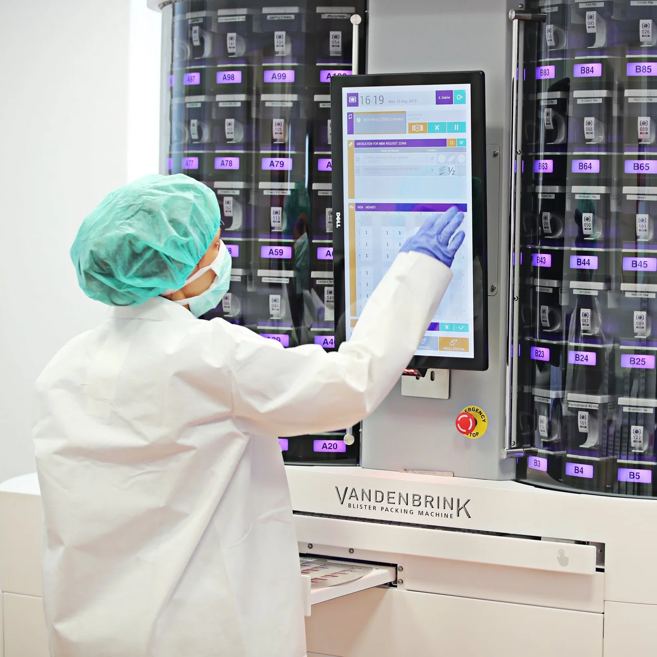 Mitarbeiterin in Schutzkleidung bedient per Touchscreen eine automatische Verblisterungsmaschine mit vielen Medikamentenkassetten.