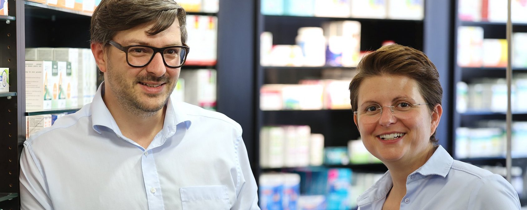 Christian und Franziska Scharpf stehen vor Regalen mit Medikamentenpackungen und halten jeweils eine Packung in der Hand; beide lächeln in die Kamera.