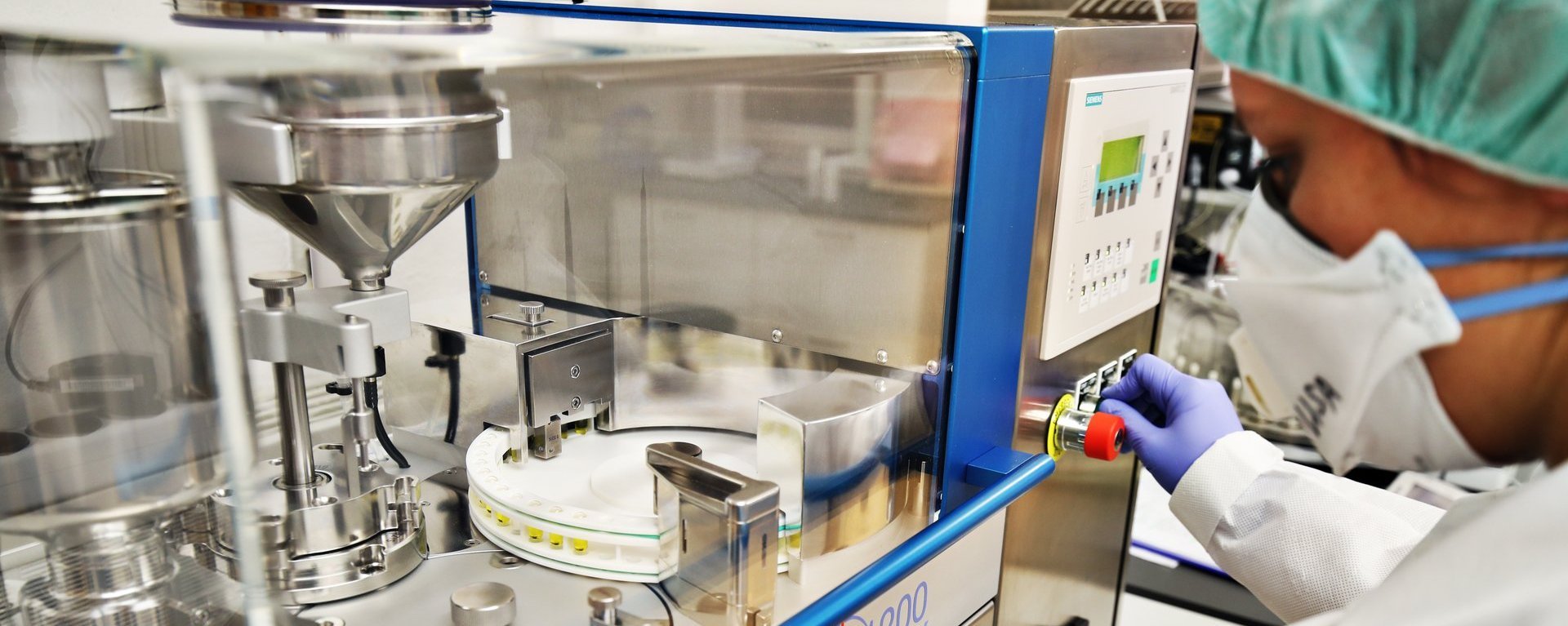 Mitarbeiterin mit Haube, Maske und Handschuhen bedient eine Verblisterungsmaschine und hält einen transparenten Behälter für Tabletten bereit.