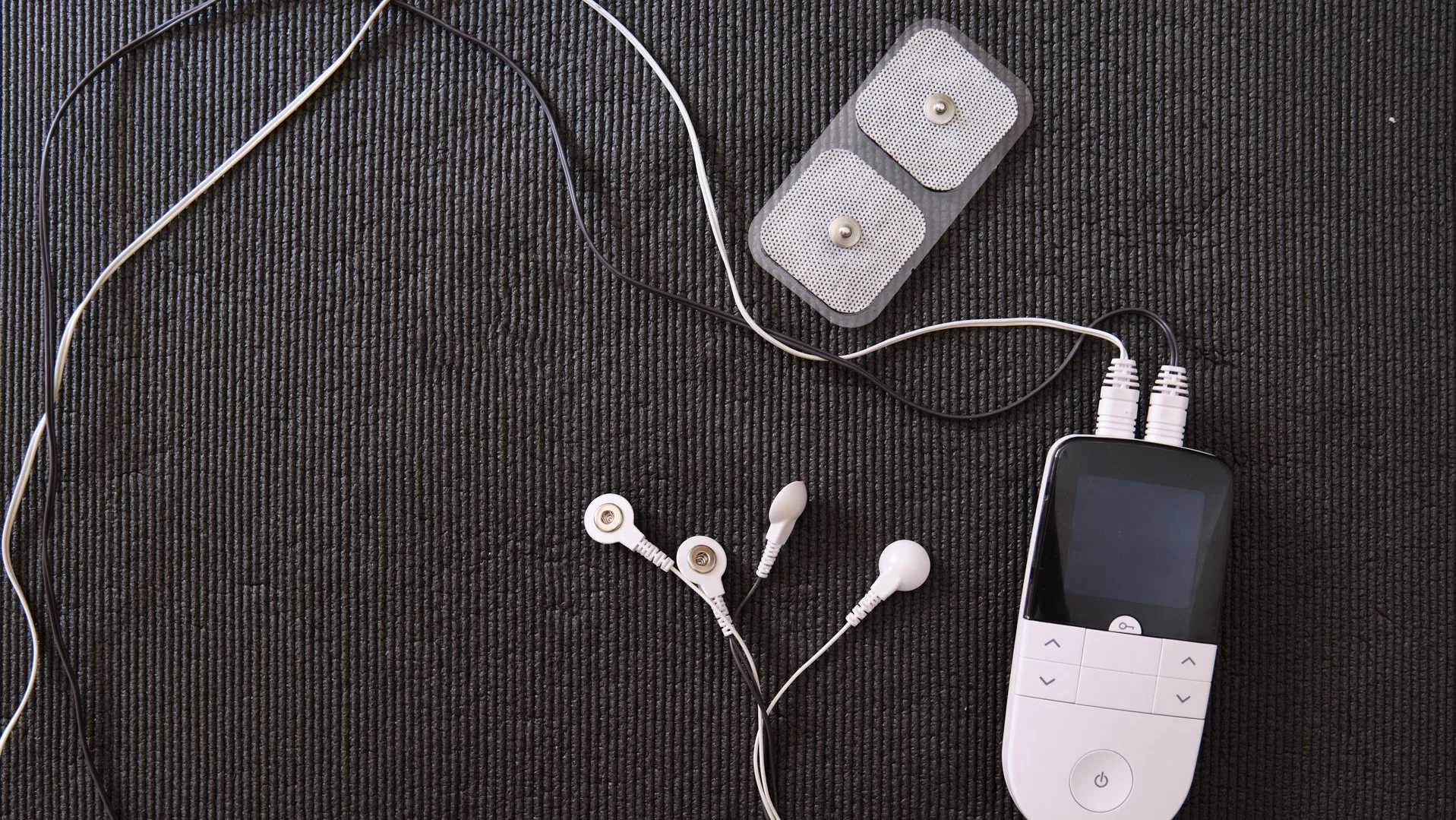 TENS/EMS-Gerät mit Bedienteil, Kabeln und Elektrodenpads liegt auf einer dunklen Unterlage.