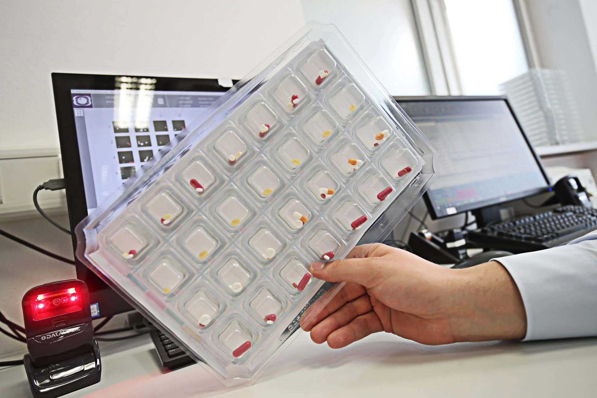 Hand hält einen Wochenblister mit einzeln verpackten Tabletten; im Hintergrund stehen Computer und Arbeitsgeräte im Apothekenbereich.