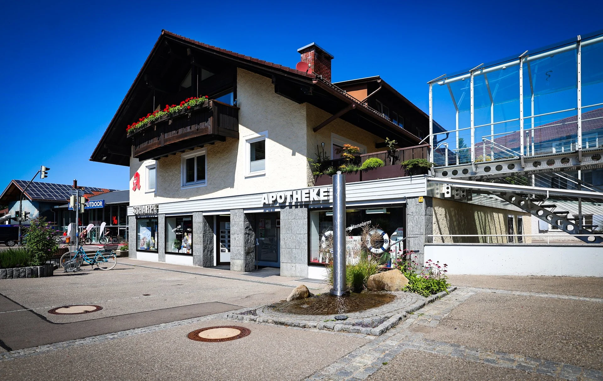 Außenansicht der Apotheke Scharpf in Sonthofen mit Eingang und Schriftzug „Apotheke“ an der Fassade; davor liegt ein kleiner Platz bei blauem Himmel.