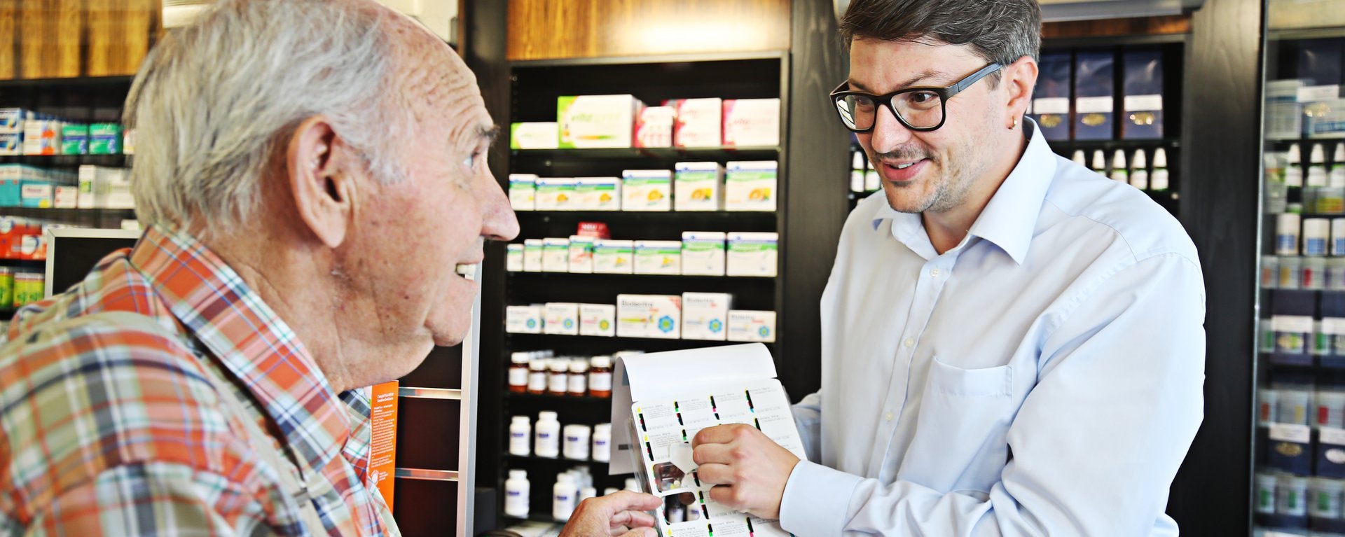 Apothekenmitarbeiter erklärt einem älteren Kunden eine Wochenblister-Verpackung mit nach Einnahmezeiten sortierten Medikamenten.
