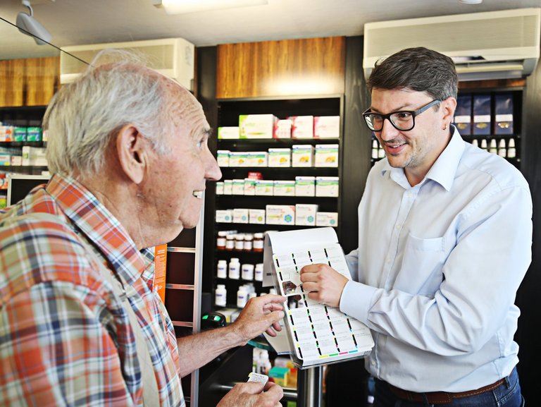 Apothekenmitarbeiter erklärt einem älteren Kunden eine Wochenblister-Verpackung mit nach Einnahmezeiten sortierten Medikamenten.