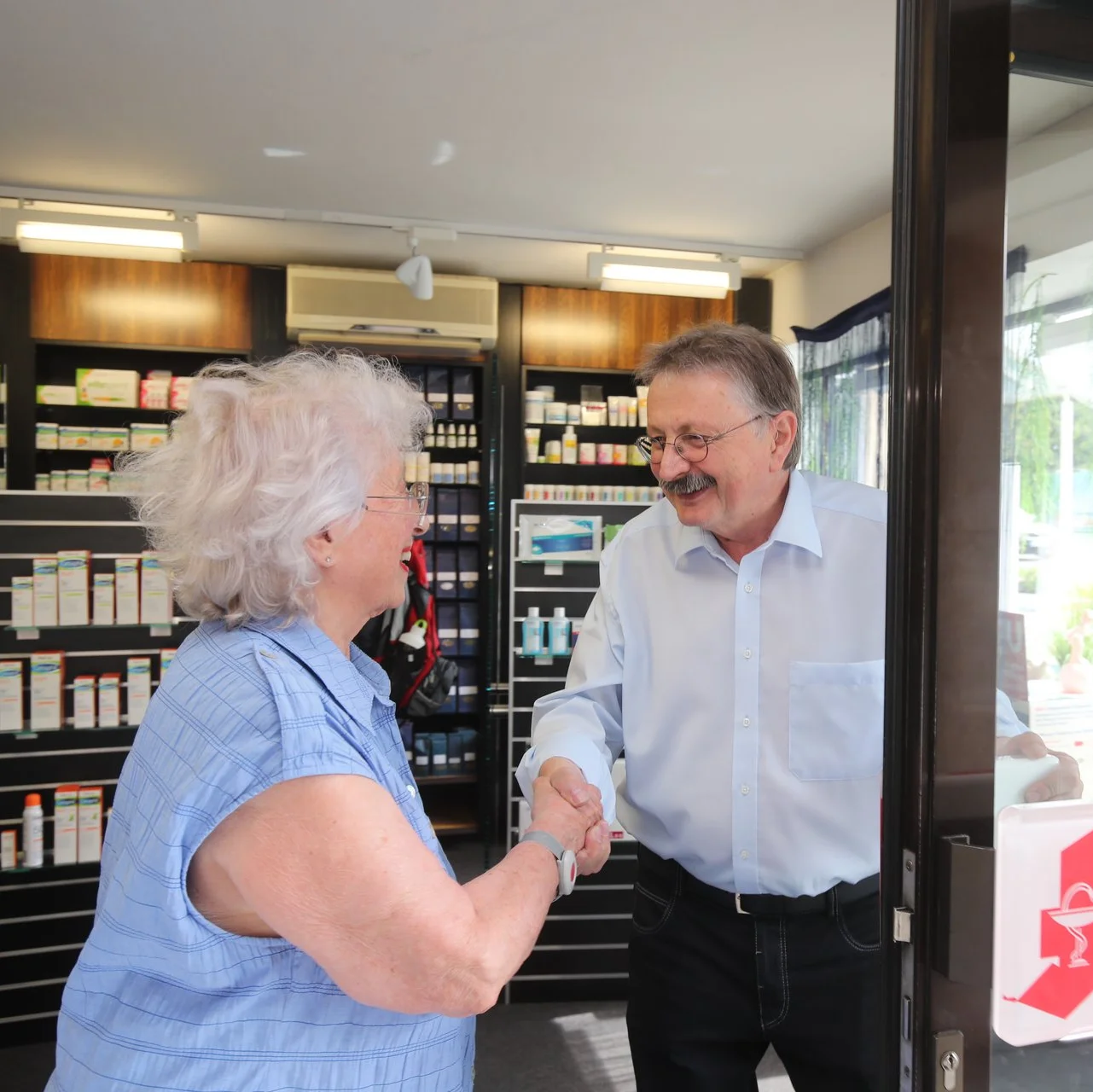 Ein Apotheker begrüßt eine Kundin im Eingangsbereich der Apotheke mit einem Handschlag; im Hintergrund sind Arzneimittelregale zu sehen.