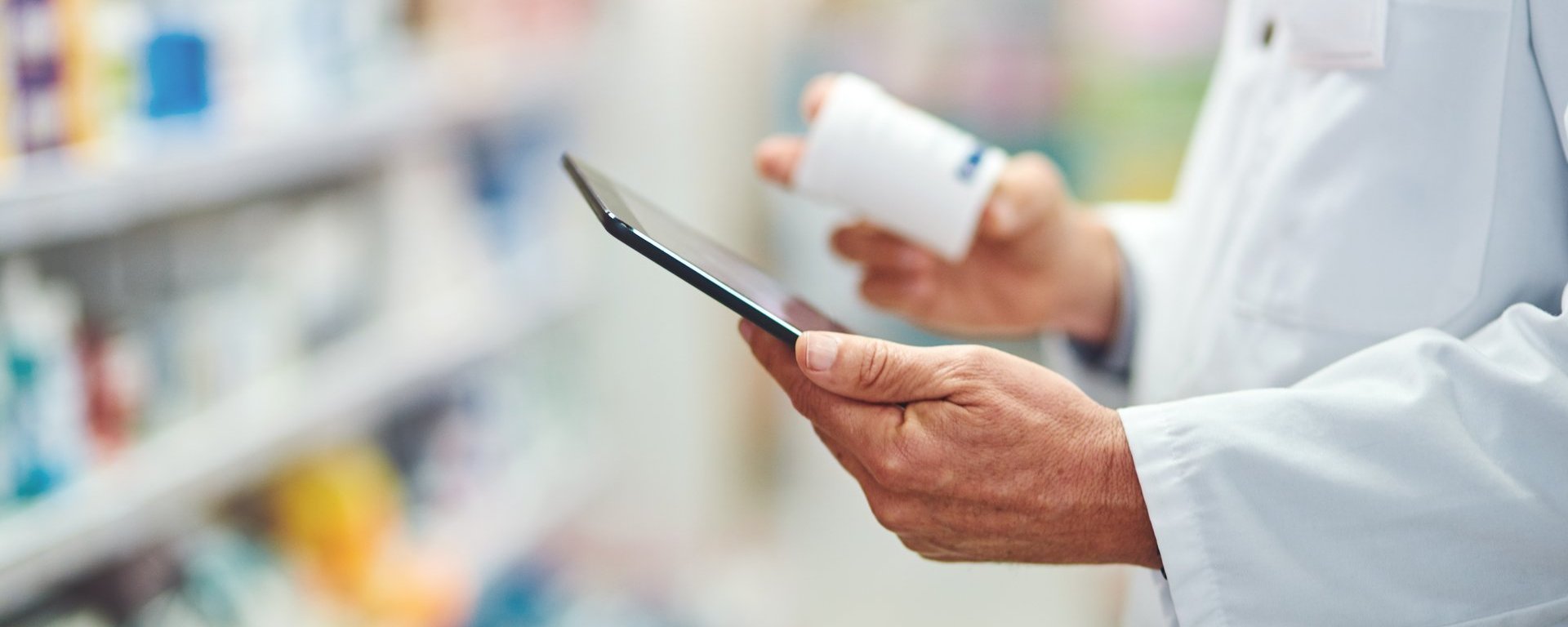 Apothekenmitarbeiter im Kittel hält ein Tablett und eine Medikamentendose in der Hand; im Hintergrund sind unscharfe Regale mit Produkten zu sehen.