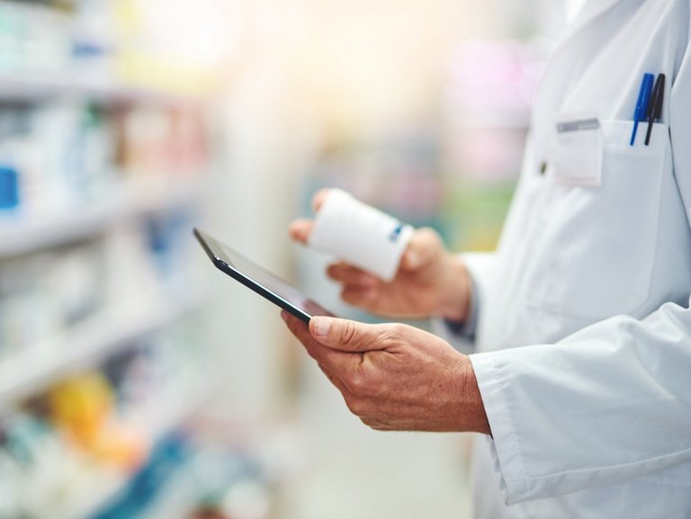 Apothekenmitarbeiter im Kittel hält ein Tablett und eine Medikamentendose in der Hand; im Hintergrund sind unscharfe Regale mit Produkten zu sehen.