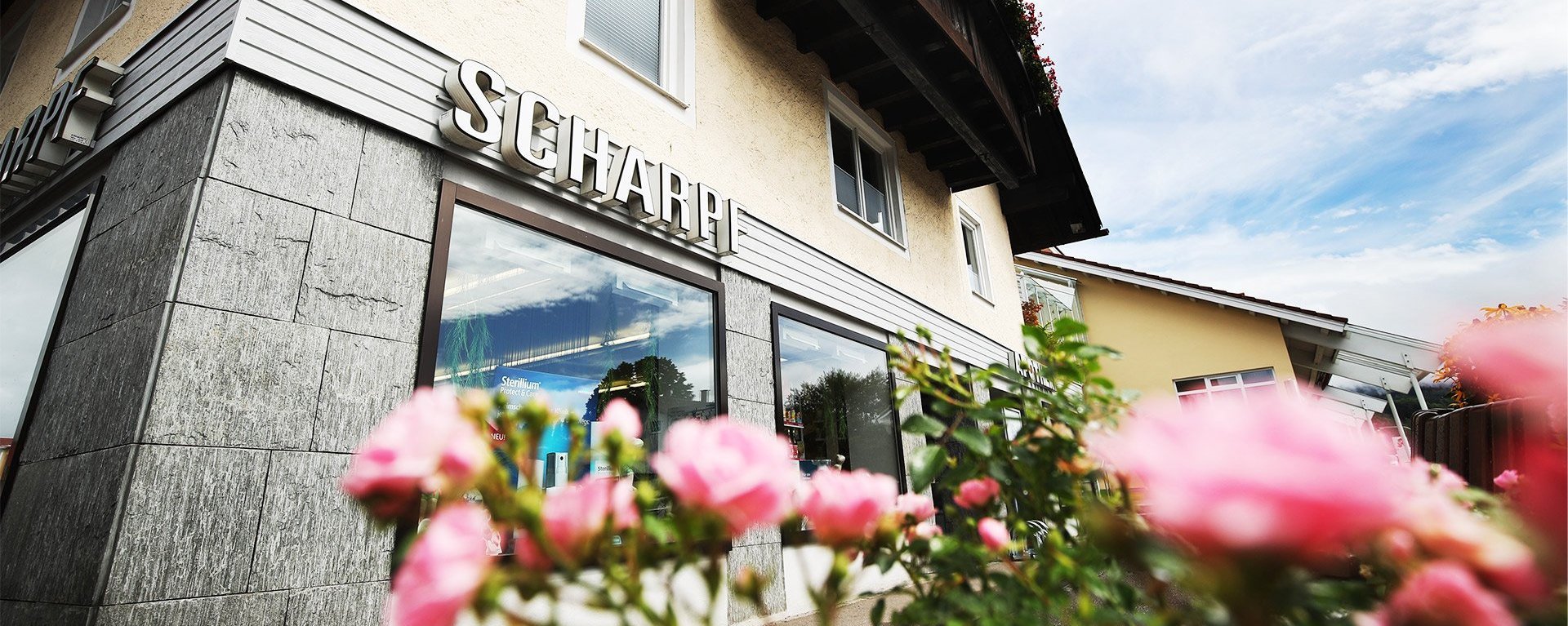 Außenansicht der Apotheke Scharpf mit Schriftzug „Scharpf“ und rotem Apotheken-A; im Vordergrund sind rosa Blumen unscharf zu sehen.