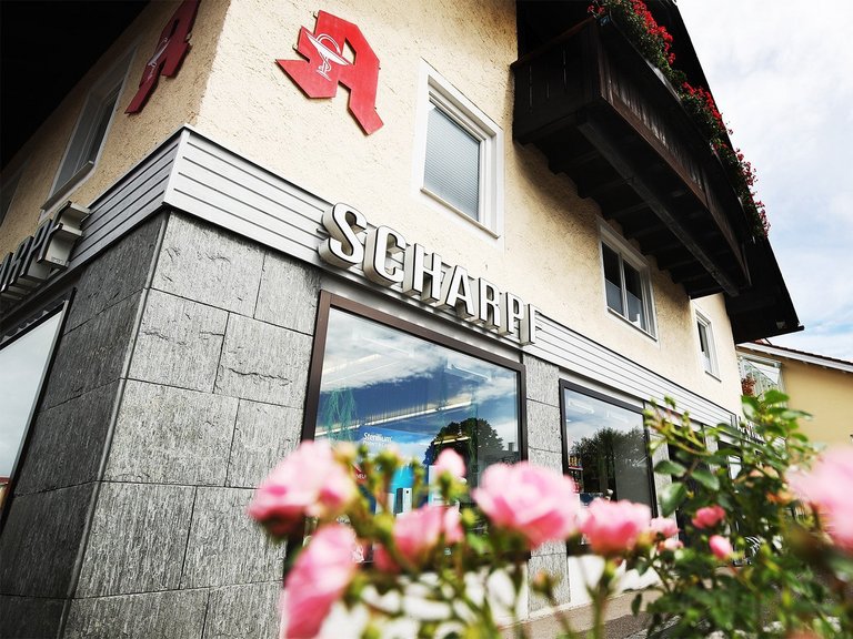 Außenansicht der Apotheke Scharpf mit Schriftzug „Scharpf“ und rotem Apotheken-A; im Vordergrund sind rosa Blumen unscharf zu sehen.