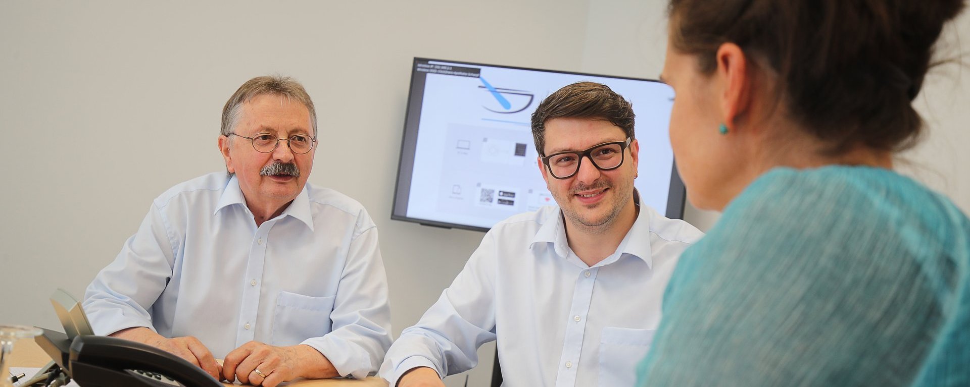 Besprechung am Tisch: zwei Männer sitzen gegenüber einer Frau und sprechen über Unterlagen; im Hintergrund hängt ein Bildschirm mit einer Präsentation.