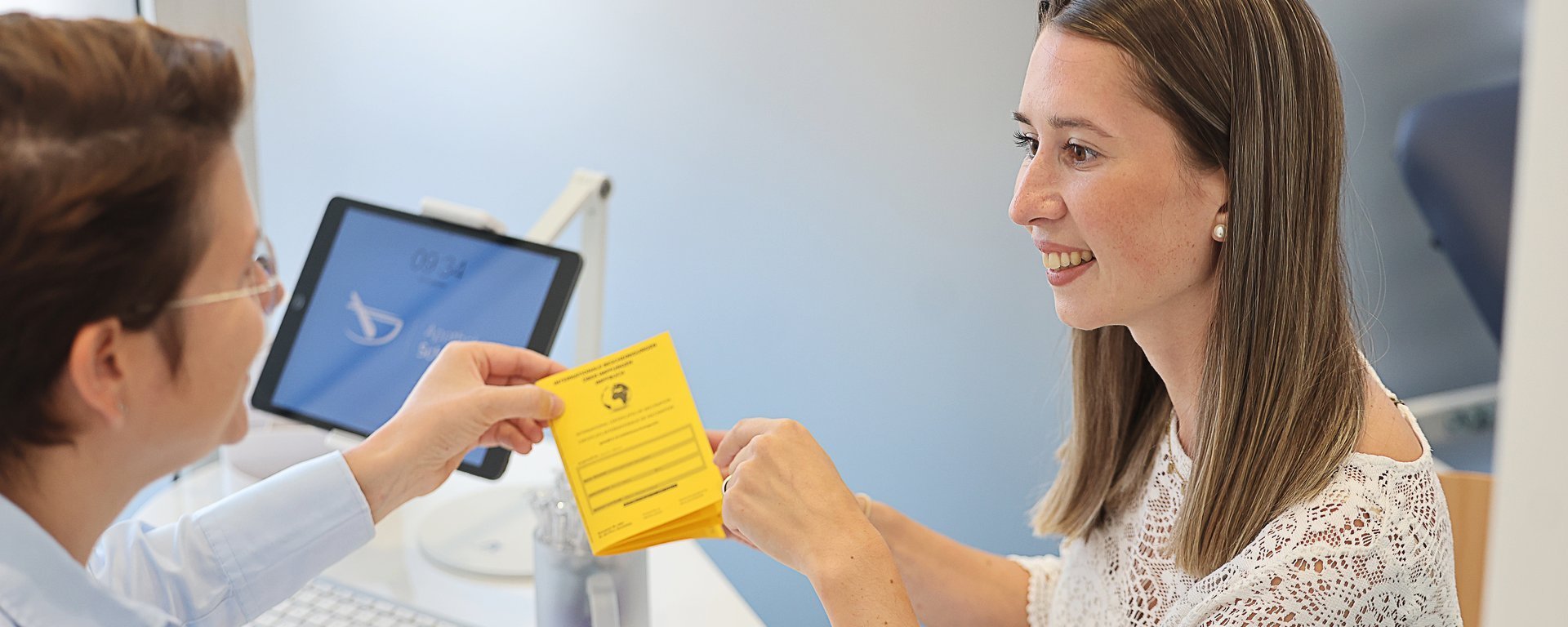 Apothekenmitarbeiterin übergibt einer Kundin am Beratungstisch ein gelbes Impfbuch/Impfdokument; beide sprechen freundlich miteinander, im Hintergrund steht ein Monitor.