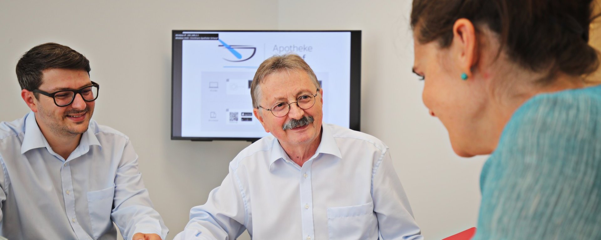 Drei Personen sitzen an einem Tisch und besprechen Unterlagen; ein Mann zeigt auf ein Dokument, im Hintergrund ist ein Bildschirm mit Präsentation zu sehen.