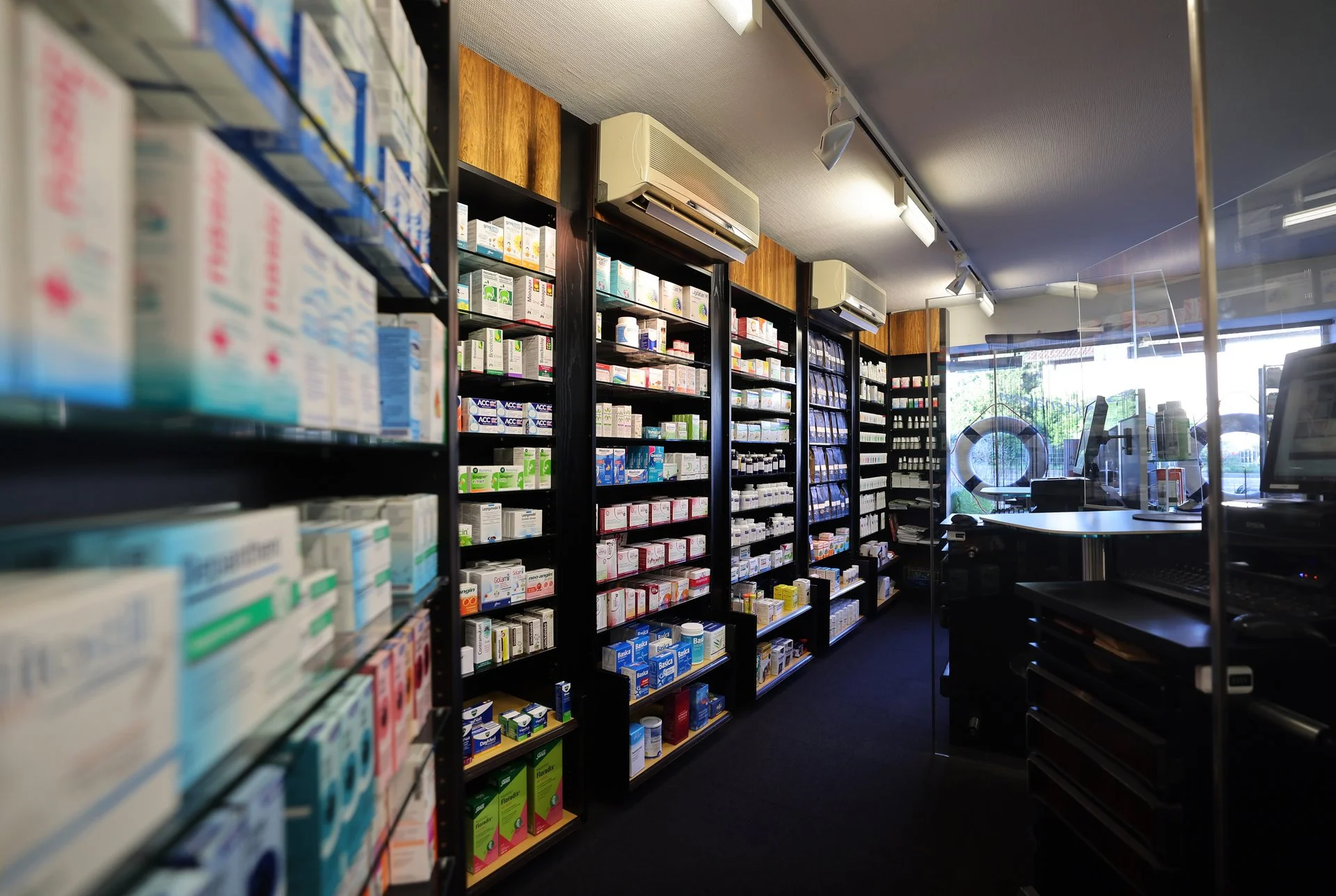 Innenansicht der Apotheke mit langen Regalen voller Arzneimittelpackungen und Blick Richtung Verkaufsbereich und Eingang.